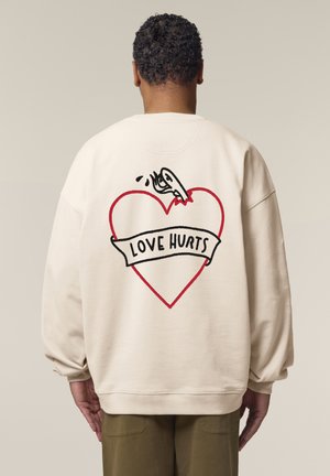 Sweatshirt surdimensionné crème avec un graphique de cœur rouge et un contour noir, ainsi que le texte "L'AMOUR FAIT MAL" sur une bannière blanche.