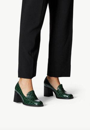 Voeten in donkergroene loafers met krokodillenpatroon en blokhak, gecombineerd met zwarte cropped broek, op een witte achtergrond.
