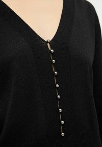 Maglione nero a V con lavorazione a maglia strutturata, dotato di sei bottoni argento lucido sul fronte, creando un sottile contrasto di materiali.