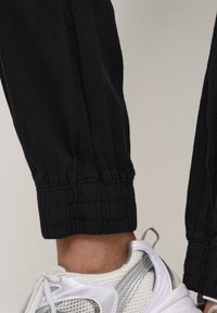 Osoba w czarnych spodniach z elastycznymi mankietami i białych, sportowych butach z siateczki oraz metaliczno-srebrnymi akcentami, stojąca na neutralnym tle.