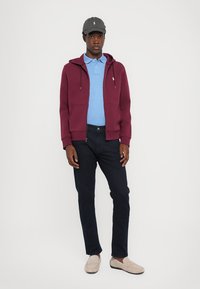 Jeune homme portant une casquette grise, un sweat à capuche bordeaux zippé, une chemise polo bleue, un jean noir et des mocassins beiges, se tenant contre un fond blanc.