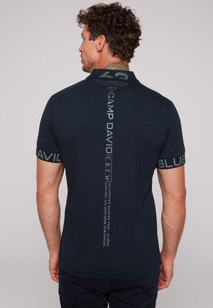 Uomo con capelli ricci e tatuaggio sul collo che indossa una polo blu navy con la scritta "Camp David" e altro testo stampato verticalmente sulla schiena.