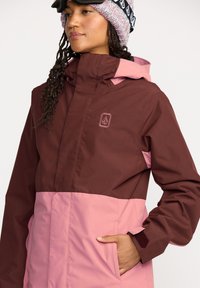 Chaqueta de invierno marrón y rosa con cremallera frontal, capucha ajustable y puños texturizados. Presenta un logotipo en el pecho y bolsillos a los lados.