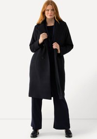 Dlouhý námořnický kabát s dvojitým zapínáním, širokými klopami a předními kapsami. Materiál se zdá být hladký s přiléhavým střihem.