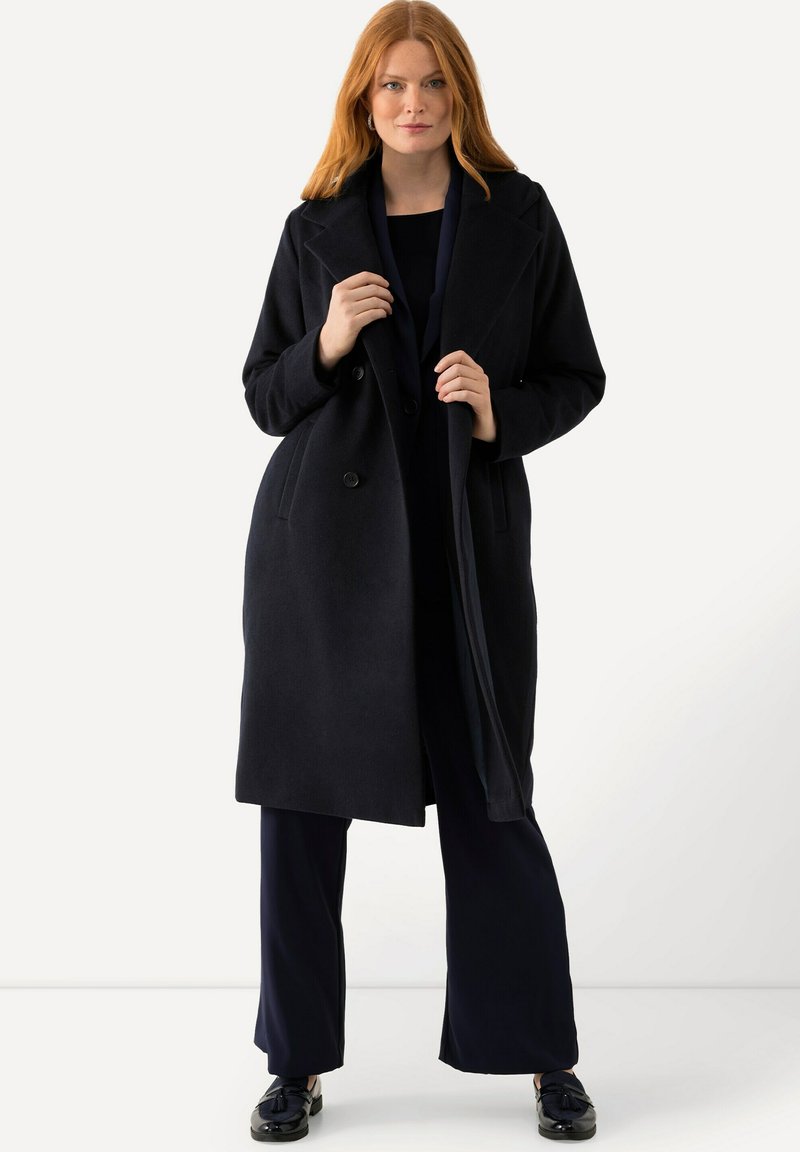 Dlouhý námořnický kabát s dvojitým zapínáním, širokými klopami a předními kapsami. Materiál se zdá být hladký s přiléhavým střihem.