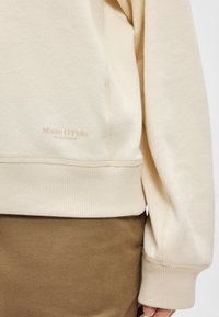 Sudadera color crema con puños y dobladillo acanalados. Presenta un sutil logo de la marca en beige claro en la parte inferior izquierda. Textura de tela suave.