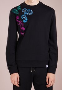 Sweat-shirt noir avec des motifs floraux brodés multicolores en vert, bleu et violet sur l'épaule gauche. Conception classique à col rond.