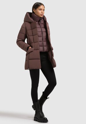 Woman wearing layered brown padded jackets, black leggings, and black chunky boots, standing with hands in jacket pockets.