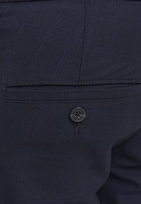 Dunkelblauer Stoff mit einem dezenten Karomuster, verfügt über eine Gesäßtasche mit einem einzelnen schwarzen Knopf und genähten Kanten.