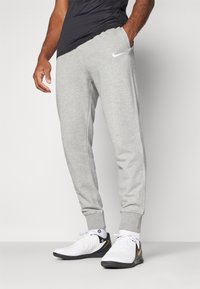 Graue Sweatpants mit schmal zulaufenden Bündchen, seitlichen Taschen und weißem Nike-Logo auf der linken Hüfte, kombiniert mit weißen Sportschuhen mit schwarzen und goldenen Akzenten.