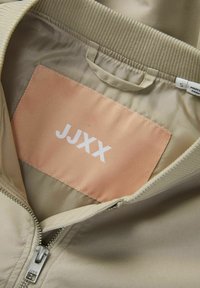 Beige jasje interieur met een licht perzikkleurig label waarop de tekst "JJXX" in het wit staat. Voorzien van een metalen rits en een geribde kraag.
