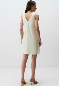 Abito senza maniche color crema con design a V sulla schiena, texture liscia e vestibilità morbida, che termina sopra il ginocchio. Indossato con sandali beige con cinturini.
