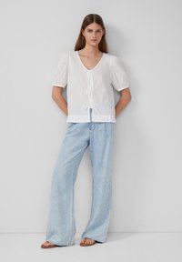 Blusa bianca con maniche a sbuffo, scollo a V e dettaglio annodato sul davanti, abbinata a jeans a gamba larga di un blu chiaro. Tessuto morbido e vestibilità rilassata.