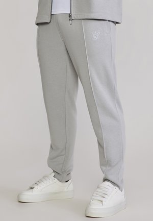 Jasnoszare spodnie dresowe z białym haftowanym logo na biodrze, w połączeniu z białymi trampkami i dopasowaną kurtką zapinaną na suwak.