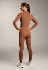 Maglietta a maniche lunghe marrone e leggings coordinati realizzati in tessuto elasticizzato, con un design aderente, colletto alto e senza marchi visibili.
