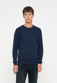 Pull en maille torsadée bleu marine avec des manches longues, un col rond et des poignets côtelés. Le matériau semble doux avec une finition texturée.