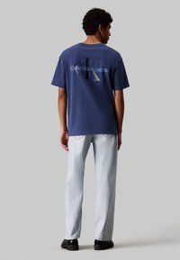 Camiseta de algodón azul marino con un gran logo "CK" en relieve, combinada con jeans azul claros y zapatos negros de suela gruesa.
