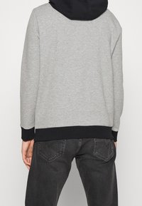 Felpa grigia con cappuccio nero, polsini e orlo a coste, presenta una texture morbida e una vestibilità rilassata. Indossata con jeans blu scuro.