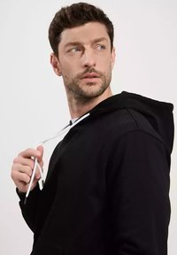 Czarna bluza z kapturem, posiadająca przednią kieszeń, regulowane białe sznurki w kapturze oraz luźny krój. Miękka struktura materiału; model patrzy w bok.