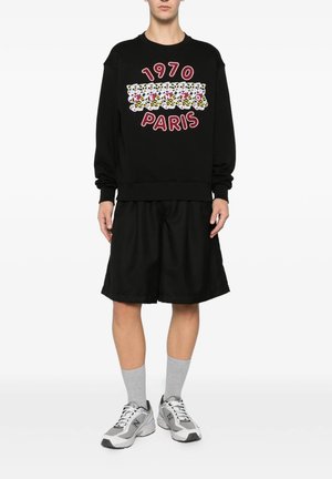 Person iført sort sweatshirt med "1970 Paris" og grafisk design, sorte vide shorts, grå sokker og grå og hvide sneakers.