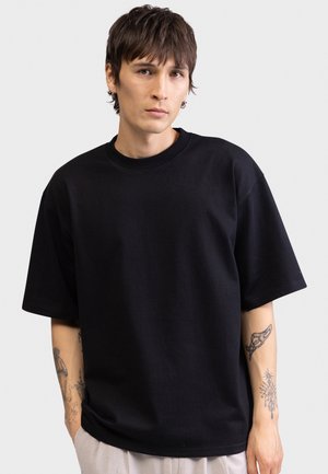 Νεαρό άτομο με κοντά καστανά μαλλιά, που φοράει μια φαρδιά μαύρη μπλούζα και ανοιχτόχρωμο παντελόνι, στέκεται μπροστά σε απλό λευκό φόντο.