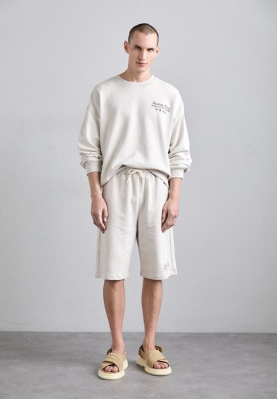 Beige bomull crew sweatshirt med logo, matchende shorts med snøring, og brune sandaler. Enkelt design og avslappet passform med myk tekstur.