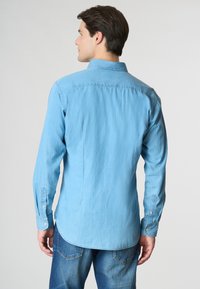 Camicia in denim azzurro chiaro con maniche lunghe, polsini con bottone e colletto. Presenta una linea dritta e una texture liscia.