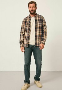 Karierte Flanellhemd in Beige, Blau und Schwarz; kombiniert mit einem cremefarbenen T-Shirt und dunklen Jeans; hellbeige Sneakers; lockere Passform.