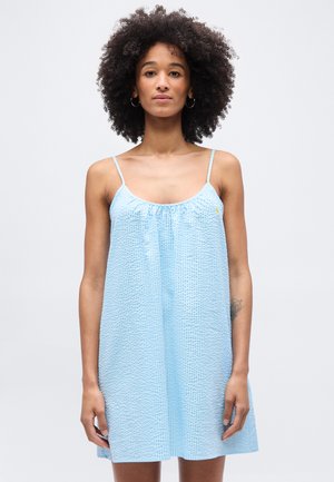 Νέα γυναίκα με φυσικά σγουρά μαλλιά που φοράει αμάνικο γαλάζιο φόρεμα με υφή, στέκεται μπροστά σε απλό ανοιχτό φόντο.