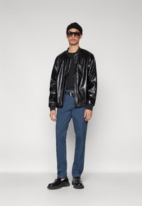 Veste bomber noire à motif crocodile, haut noir ajusté, pantalon bleu, chaussures noires et lunettes de soleil oversize. Le modèle porte un bonnet noir.