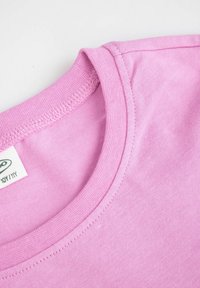 Zbliżenie różowej bawełnianej koszulki z okrągłym dekoltem, ukazujące szczegółowe szwy oraz częściową metkę na białym tle.