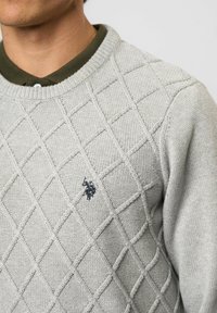 Muž nosiaci svetlosivý sveter s diamantovým vzorom cez olivovo zelenú košeľu s golierom a malým čiernym vyšívaným logom na hrudi.