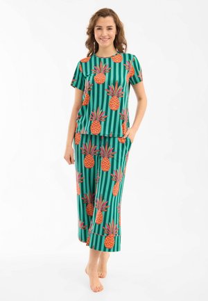 Donna sorridente mentre è in piedi a piedi nudi in pigiama verde con righe verticali e grandi motivi di ananas arancione.