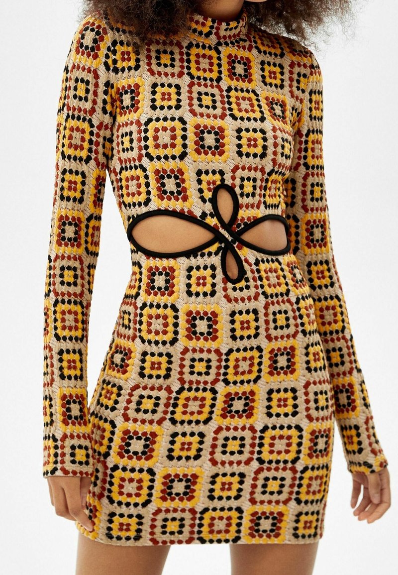 Robe courte en maille à manches longues avec motif géométrique en jaune, orange, rouge et noir, présentant une découpe en forme de rosace avec bordure noire à la taille.