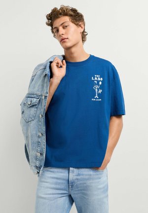 Junger Mann trägt ein blaues "The Lads Pepe Jeans" T-Shirt, hält eine Jeansjacke über einer Schulter und steht vor einem einfarbigen Hintergrund.