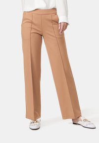 Pantalon ample couleur tan en tissu lisse avec des plis à l'avant. Porté avec des chaussures blanches sans lacets ornées d'accents dorés.