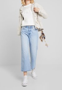 Hellgraue Jacke mit Kordelzügen, weißer Strickpullover mit strukturiertem Muster, hochgeschnittene hellblaue Jeans und weiße Stoffsportchuhe.