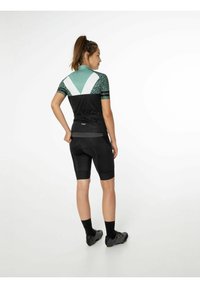 Korte mouwen fietsshirt in zwart, teal en wit met een gepatternde schouderpartij. Het shirt is gecombineerd met zwarte gevoerde shorts en fietsschoenen.