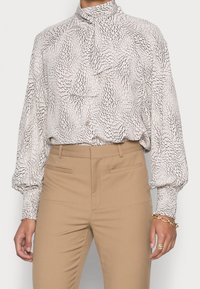 Blouse à manches longues avec un col haut, en tissu texturé crème avec un motif abstrait noir, associée à un pantalon beige ajusté.