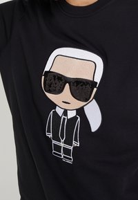 Personnage de dessin animé avec cheveux blancs en queue de cheval, lunettes de soleil noires, costume noir et cravate brodée sur tissu noir.