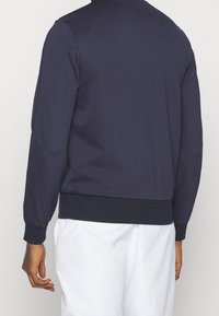 Marineblaue Sweatjacke mit strukturiertem Stoff, gerippten Bündchen und Saum, Rundhalsausschnitt und körpernahem Schnitt, getragen mit weißen Shorts.