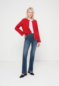 Κόκκινη πλεκτή ζακέτα με φθαρμένα άκρα, φορεμένη πάνω από λευκό τοπ. Συνδυασμένη με σκουρόχρωμα τζιν καμπάνα και μαύρα παπούτσια με μυτερή μύτη.