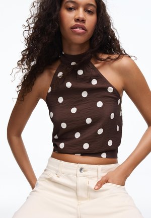 Ženska s kodrastimi lasmi, oblečena v rjavo brezrokavnico z belimi pikami in kremasto bele hlače z visokim pasom.