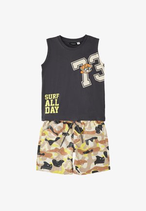Top sin mangas gris oscuro con "73," un oso con sombrero y el texto "SURF ALL DAY", combinado con pantalones cortos de camuflaje en amarillo, marrón y negro.