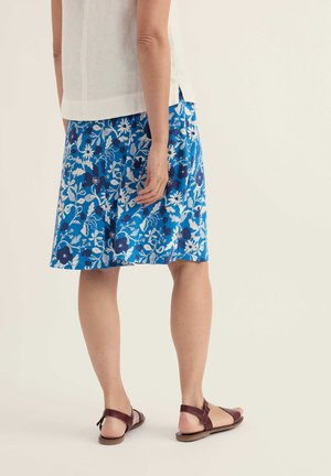 Naine, kes kannab sinist lillelist põlvpikkust seelikut, valget varrukateta toppi ja pruunid lamedad sandaale, seisab tavalise tausta ees.