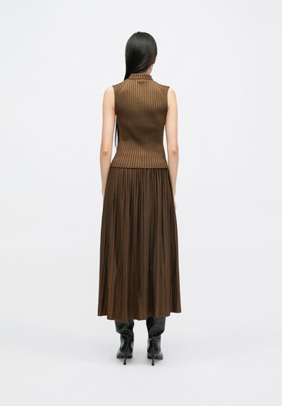 Brown ribbed sleeveless top with a high collar, paired with a flowing, pleated ankle-length skirt. Worn with black knee-high boots.