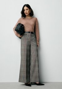 Sieviete ģērbta brūnā džemperī, ar augsto jostasvietu, platu svītrainu biksēm, melnām augstpapēžu kurpēm un tur rokās melnu ādas rokassomu, stāv pretī vienkāršai sienai.