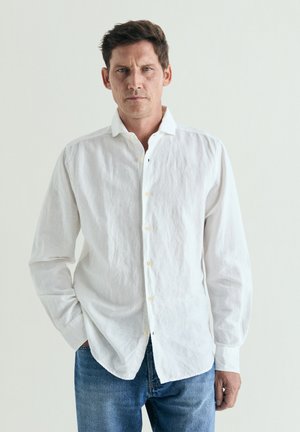 Hombre con cabello corto y oscuro, vistiendo una camisa blanca abotonada y jeans azules, de pie contra un fondo claro y liso.