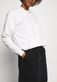 White hooded sweatshirt with a front pocket and drawstrings, paired with a black skirt featuring white stitching details. Textured fabric.