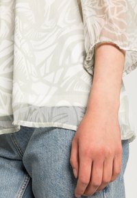 Blusa leve e transparente com um padrão de folhas em verde-claro e branco. Apresenta mangas franzidas e uma bainha que termina acima da cintura.